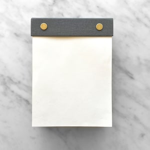 May include: A blank white notepad with a grey fabric cover and two gold metal push pins. The notepad is on a white marble surface.