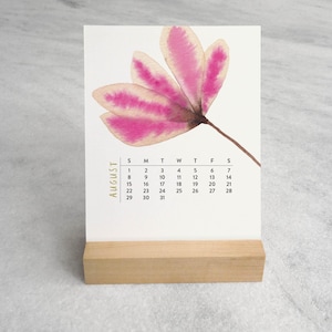 Peut inclure: Un calendrier de bureau pr&eacute;sentant une illustration &agrave; l'aquarelle d'une fleur rose &agrave; cinq p&eacute;tales. La page du calendrier affiche le mois d'ao&ucirc;t avec les jours de la semaine et les dates sous la fleur. Il est pos&eacute; sur un support en bois clair.