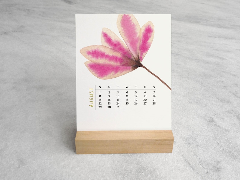 Peut inclure: Un calendrier de bureau pr&eacute;sentant une illustration &agrave; l'aquarelle d'une fleur rose &agrave; cinq p&eacute;tales. La page du calendrier affiche le mois d'ao&ucirc;t avec les jours de la semaine et les dates sous la fleur. Il est pos&eacute; sur un support en bois clair.