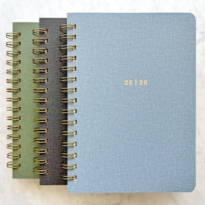 May include: Three spiral-bound notebooks in shades of green, gray, and blue. The notebooks have gold-colored spiral bindings and the blue notebook has the numbers "25 | 26" printed in gold. The covers have a textured appearance.