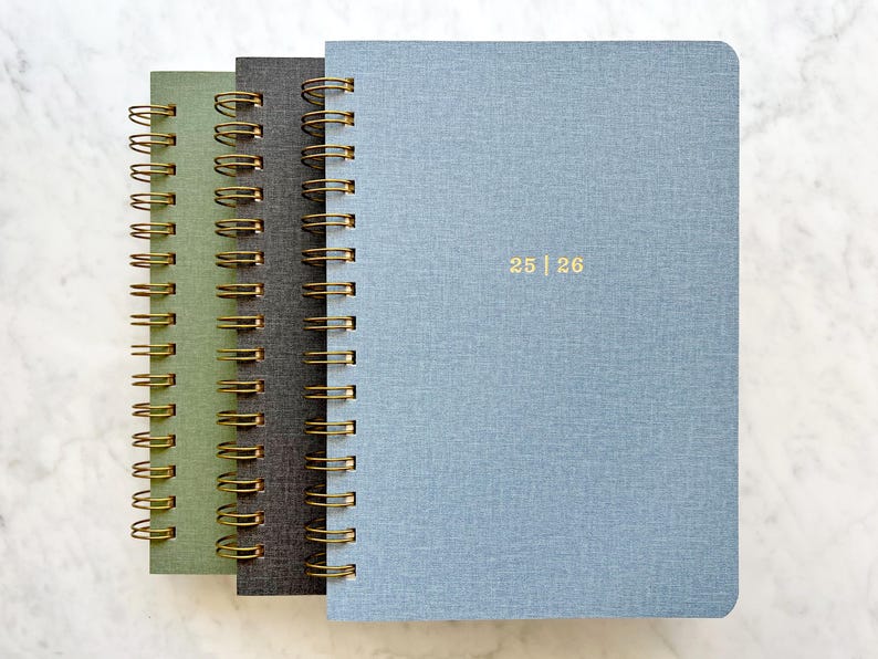 May include: Three spiral-bound notebooks in shades of green, gray, and blue. The notebooks have gold-colored spiral bindings and the blue notebook has the numbers "25 | 26" printed in gold. The covers have a textured appearance.