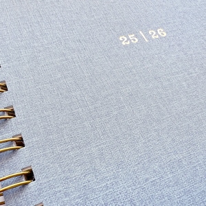 May include: Close-up of a light blue spiral-bound notebook. The cover has a textured fabric appearance with the numbers "25 | 26" printed in a light color. The spiral binding is a gold color.