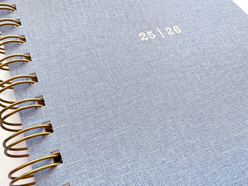 May include: Close-up of a light blue spiral-bound notebook. The cover has a textured fabric appearance with the numbers "25 | 26" printed in a light color. The spiral binding is a gold color.