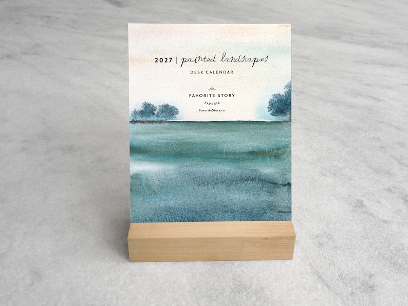 May include: A 2027 desk calendar featuring a watercolor landscape painting of a teal and blue horizon with trees. The text reads 2027 | painted landscapes DESK CALENDAR, THE FAVORITE STORY PAPERIE, FavoriteStory.co. It sits on a light wood block stand.