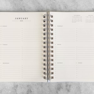 May include: Open spiral-bound planner, displaying the month of January 2025. The left page shows the days of the week with checklist boxes. The right page has a calendar and checklist boxes. The planner is open on a gray surface.