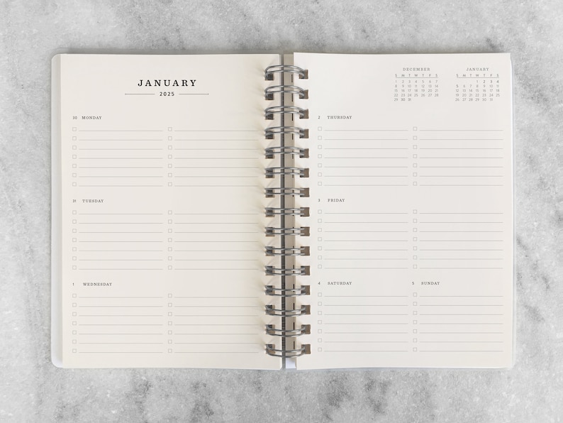 May include: Open spiral-bound planner, displaying the month of January 2025. The left page shows the days of the week with checklist boxes. The right page has a calendar and checklist boxes. The planner is open on a gray surface.