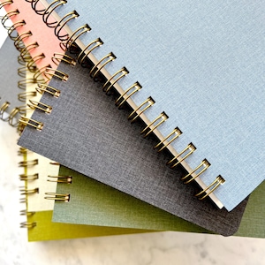 May include: A stack of spiral-bound notebooks in various colors, including pink, gray, blue, and green. The notebooks have gold-colored metal spiral bindings.