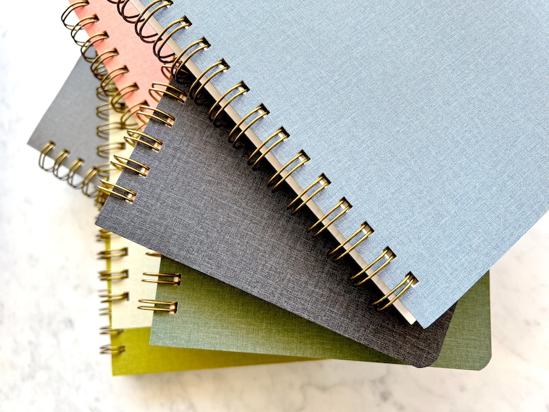 May include: A stack of spiral-bound notebooks in various colors, including pink, gray, blue, and green. The notebooks have gold-colored metal spiral bindings.