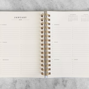 May include: An open spiral bound planner with a white cover and gold binding. The planner is open to January 2025 and has a calendar view for December 2024 and January 2025. The planner has a daily layout with space for notes and tasks.