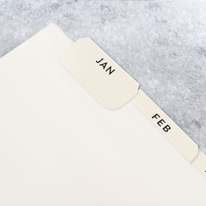 May include: Cream-colored paper tabs labeled with the months JAN, FEB, and MAR, attached to a stack of white paper. The tabs are in a serif font and are set against a gray background.