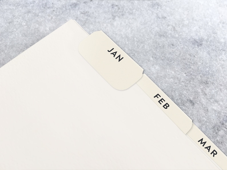 May include: Cream-colored paper tabs labeled with the months JAN, FEB, and MAR, attached to a stack of white paper. The tabs are in a serif font and are set against a gray background.