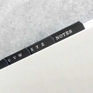May include: Close-up of a notebook with black divider tabs. The tabs are labeled with white text, including letters and the word "NOTES". The notebook pages are white, and the background is a light gray.