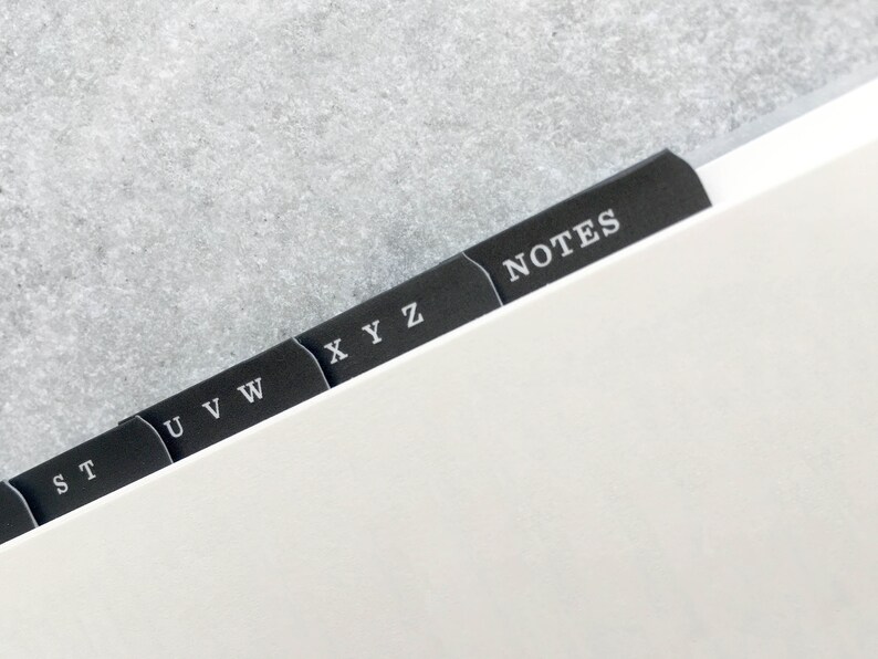May include: Close-up of a notebook with black divider tabs. The tabs are labeled with white text, including letters and the word "NOTES". The notebook pages are white, and the background is a light gray.