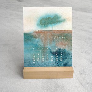 May include: A desk calendar featuring a watercolor landscape painting of trees on a cliff above water. The word January is written in white script above a grid of dates. The calendar is held upright by a rectangular light-colored wooden stand.