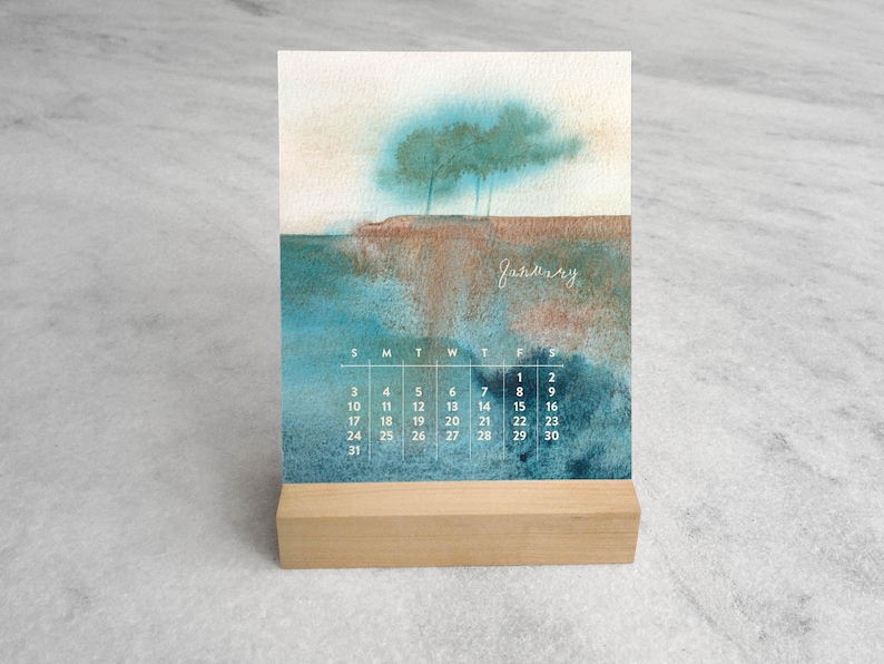 May include: A desk calendar featuring a watercolor landscape painting of trees on a cliff above water. The word January is written in white script above a grid of dates. The calendar is held upright by a rectangular light-colored wooden stand.