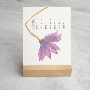Peut inclure: Un calendrier de bureau pour le mois de d&eacute;cembre pr&eacute;sentant une illustration &agrave; l'aquarelle d'une fleur violette avec une longue tige dor&eacute;e. La grille du calendrier affiche les jours du mois en noir sur fond blanc, sur un support en bois clair.