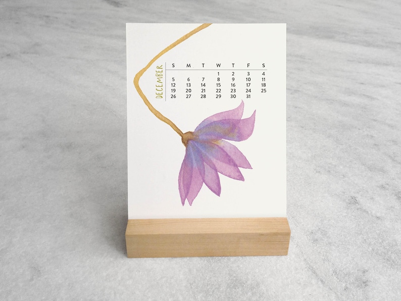 Peut inclure: Un calendrier de bureau pour le mois de d&eacute;cembre pr&eacute;sentant une illustration &agrave; l'aquarelle d'une fleur violette avec une longue tige dor&eacute;e. La grille du calendrier affiche les jours du mois en noir sur fond blanc, sur un support en bois clair.