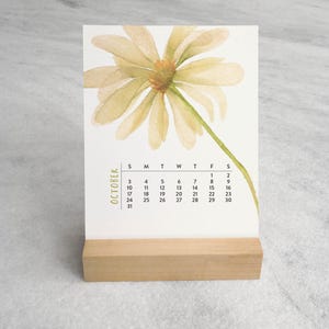 Peut inclure: Un calendrier de bureau rectangulaire pour le mois d'octobre pr&eacute;sentant une illustration &agrave; l'aquarelle d'une marguerite jaune p&acirc;le avec une tige verte. La grille du calendrier est imprim&eacute;e en noir et tenue par un support en bois clair.