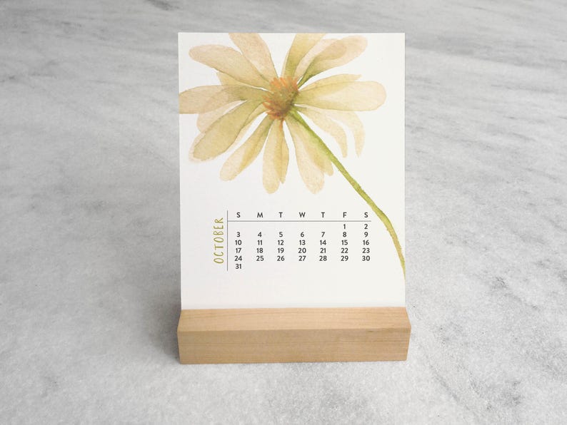 Peut inclure: Un calendrier de bureau rectangulaire pour le mois d'octobre pr&eacute;sentant une illustration &agrave; l'aquarelle d'une marguerite jaune p&acirc;le avec une tige verte. La grille du calendrier est imprim&eacute;e en noir et tenue par un support en bois clair.