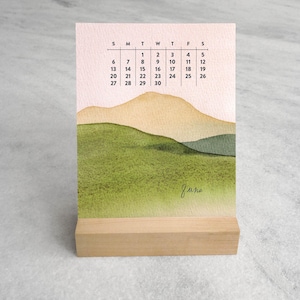 May include: A vertical desk calendar for the month of June featuring a watercolor landscape of green hills and tan mountains against a pale pink sky. The calendar grid displays the days of the month from 1 to 30 in black text on a white background.