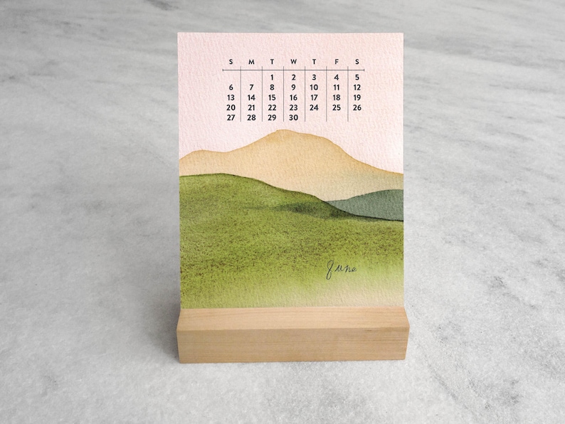 May include: A vertical desk calendar for the month of June featuring a watercolor landscape of green hills and tan mountains against a pale pink sky. The calendar grid displays the days of the month from 1 to 30 in black text on a white background.