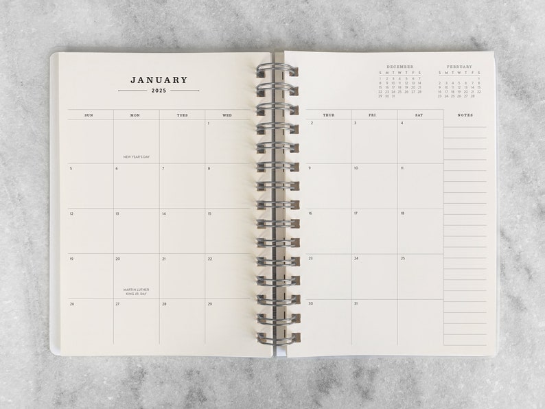 May include: Open spiral-bound planner, displaying the January 2025 calendar. The cream-colored pages feature a monthly layout with individual day boxes and a notes section. The calendar includes New Year's Day and Martin Luther King Jr. Day.