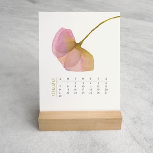 Peut inclure: Une carte de calendrier de bureau blanche avec une illustration &agrave; l'aquarelle d'une fleur rose unique avec une tige verte. Le texte FEBRUARY est imprim&eacute; verticalement en dor&eacute;, suivi d'une grille noire des dates du mois, sur un support en bois.