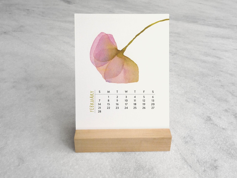 Peut inclure: Une carte de calendrier de bureau blanche avec une illustration &agrave; l'aquarelle d'une fleur rose unique avec une tige verte. Le texte FEBRUARY est imprim&eacute; verticalement en dor&eacute;, suivi d'une grille noire des dates du mois, sur un support en bois.