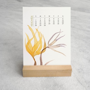 Peut inclure: Un calendrier de bureau vertical pour novembre avec une illustration &agrave; l'aquarelle d'&eacute;l&eacute;ments botaniques jaunes et bruns. Le calendrier est maintenu droit par un support en bloc de bois clair rectangulaire. Le texte November est imprim&eacute; verticalement.