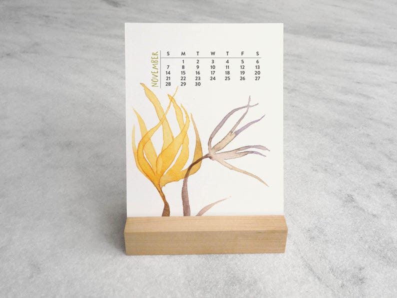 Peut inclure: Un calendrier de bureau vertical pour novembre avec une illustration &agrave; l'aquarelle d'&eacute;l&eacute;ments botaniques jaunes et bruns. Le calendrier est maintenu droit par un support en bloc de bois clair rectangulaire. Le texte November est imprim&eacute; verticalement.