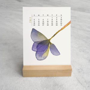 Peut inclure: Une page de calendrier de bureau verticale pour juillet avec une illustration &agrave; l'aquarelle d'une fleur violette et jaune. La grille du calendrier affiche les jours du mois en noir, le tout maintenu par un support en bois rectangulaire.