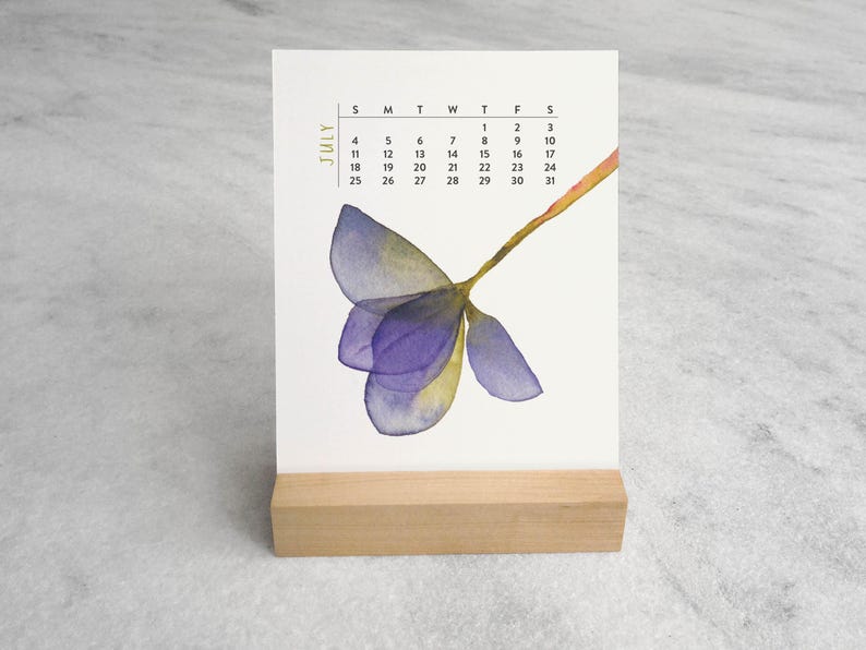 Peut inclure: Une page de calendrier de bureau verticale pour juillet avec une illustration &agrave; l'aquarelle d'une fleur violette et jaune. La grille du calendrier affiche les jours du mois en noir, le tout maintenu par un support en bois rectangulaire.