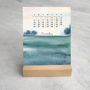 May include: A desk calendar for December featuring a watercolor landscape of a teal and blue sea with distant trees. The calendar grid displays the days of the month in black text above the word December written in a cursive script font.