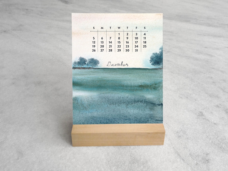 May include: A desk calendar for December featuring a watercolor landscape of a teal and blue sea with distant trees. The calendar grid displays the days of the month in black text above the word December written in a cursive script font.