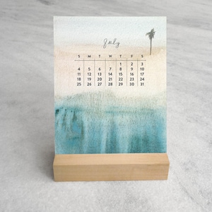 May include: A desk calendar for July featuring a watercolor illustration of a sandy beach and blue ocean with a single palm tree. The calendar grid displays the days of the month in black text, all held upright by a rectangular light wood base.