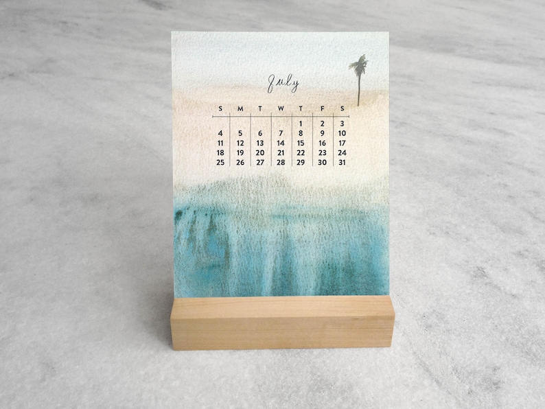 May include: A desk calendar for July featuring a watercolor illustration of a sandy beach and blue ocean with a single palm tree. The calendar grid displays the days of the month in black text, all held upright by a rectangular light wood base.