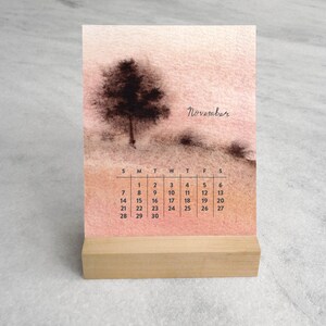 May include: A desk calendar for November featuring a watercolor-style illustration of a dark tree on a soft peach and tan background. The calendar grid displays the days of the month in black text, and the entire piece rests on a light wood base.