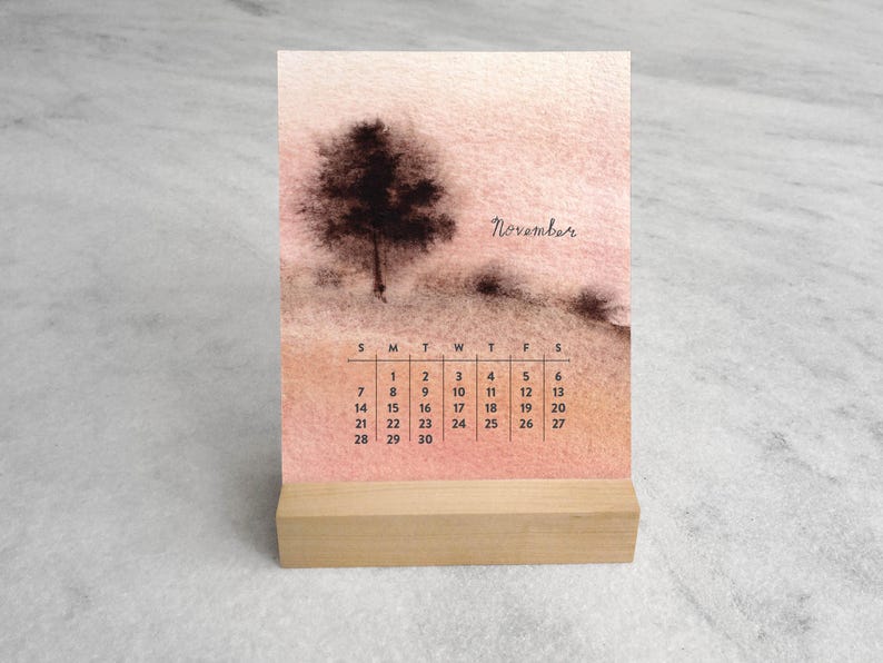 May include: A desk calendar for November featuring a watercolor-style illustration of a dark tree on a soft peach and tan background. The calendar grid displays the days of the month in black text, and the entire piece rests on a light wood base.