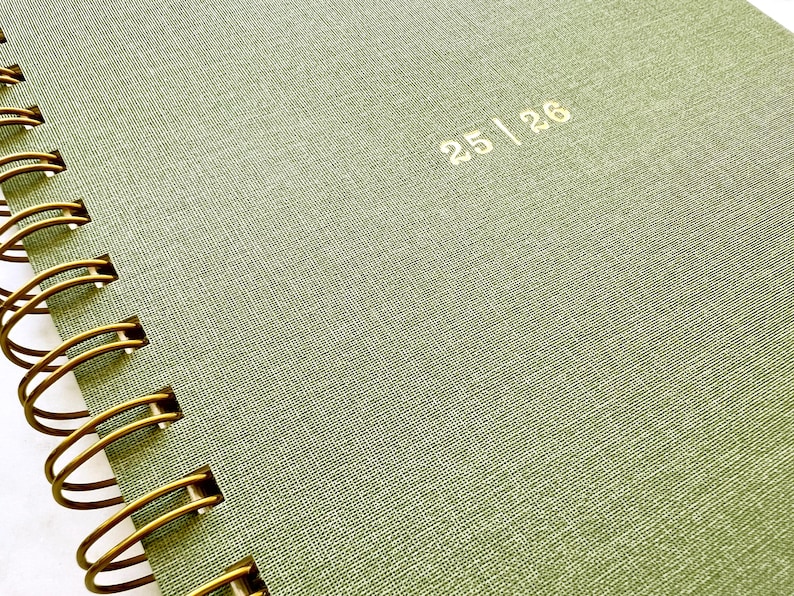 May include: Close-up of a spiral-bound notebook with a textured, olive-green cover. The gold spiral binding is visible along the left edge. The numbers "25 | 26" are printed in gold on the cover.