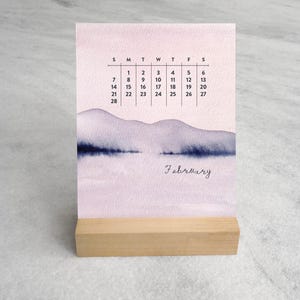 May include: A desk calendar for February featuring a watercolor illustration of purple mountains and a dark blue horizon line. The calendar displays the days of the month in a grid format with black text, all supported by a light-colored wooden base.
