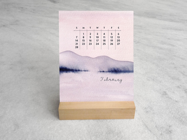 May include: A desk calendar for February featuring a watercolor illustration of purple mountains and a dark blue horizon line. The calendar displays the days of the month in a grid format with black text, all supported by a light-colored wooden base.