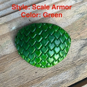 May include: Green scale armour piece. The piece is oval-shaped and features a textured pattern of overlapping scales. The text "Style: Scale Armor Color: Green" is visible in the image.