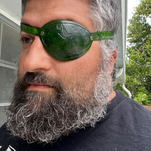 May include: A green leather eye patch with a dragonfly design. The eye patch is secured with a strap and metal rivets. The eye patch is designed to cover the left eye.