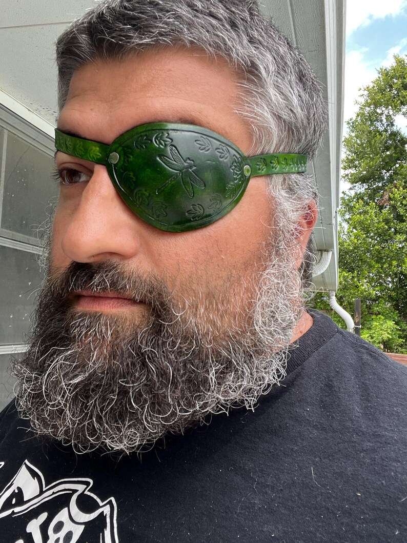 May include: A green leather eye patch with a dragonfly design. The eye patch is secured with a strap and metal rivets. The eye patch is designed to cover the left eye.