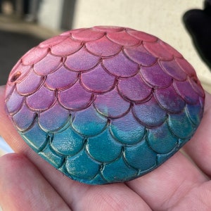 May include: A round, hand-painted object with a mermaid scale design. The colors transition from pink to purple to teal. The object is held in a hand.