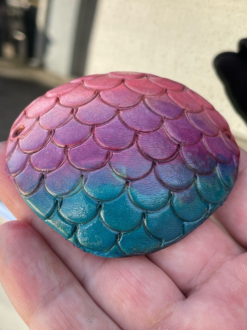 May include: A round, hand-painted object with a mermaid scale design. The colors transition from pink to purple to teal. The object is held in a hand.