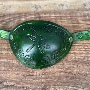 May include: A handcrafted, dark green leather eye patch. The eye patch features a dragonfly design in the center, surrounded by leaf accents. The strap is also green and has leaf-shaped cutouts. The eye patch is secured with metal rivets.