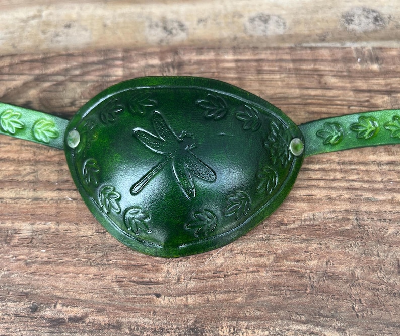 May include: A handcrafted, dark green leather eye patch. The eye patch features a dragonfly design in the center, surrounded by leaf accents. The strap is also green and has leaf-shaped cutouts. The eye patch is secured with metal rivets.
