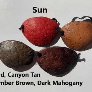 May include: Four leather eye patches in red, canyon tan, timber brown, and dark mahogany. Each patch has a sunburst design and a black elastic band. The word "Sun" is above the patches.