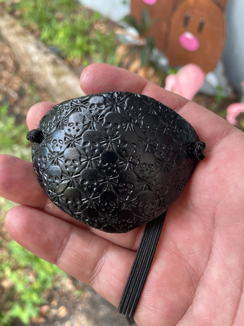 May include: Black eye patch with a skull and crossbones pattern. The eye patch is held in a hand and has a black elastic band attached for wear. The eye patch is made of a textured material.